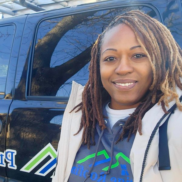 Prestige Roofing employee with two-tone locs holds a clipboard and smiles beside a dark blue service vehicle