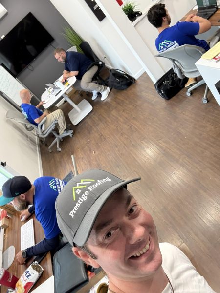 A man in a grey Prestige Roofing hat taking a selfie with team members working in a modern office